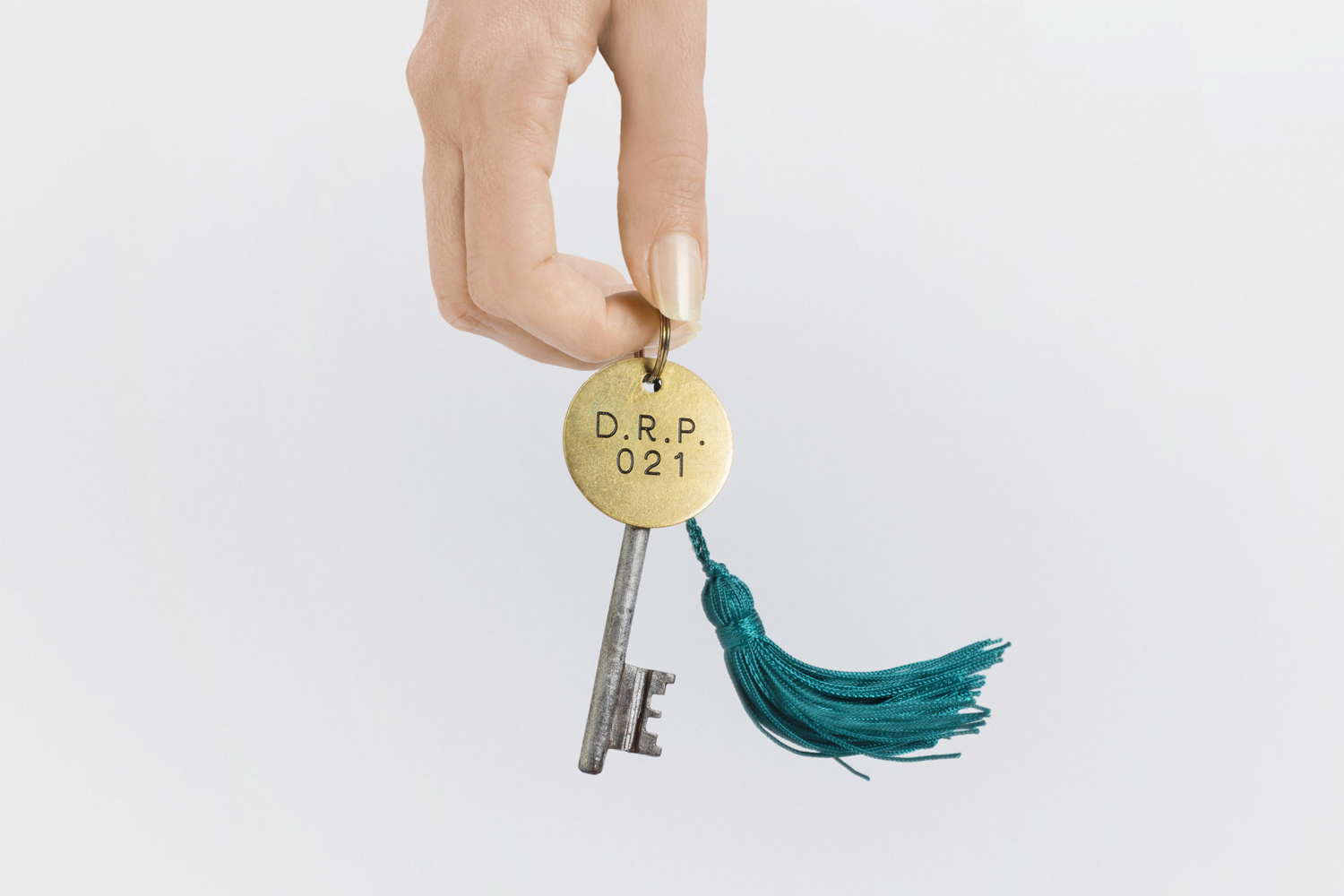 Hand holding a brass room key with a teal tassel against a plain white background