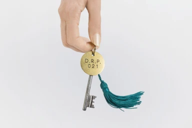 Hand holding a brass room key with a teal tassel against a plain white background