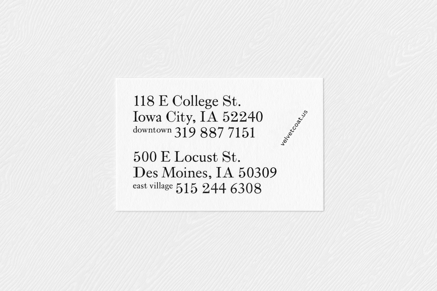 White business card with black typography displaying addresses for Velvet Coat, resting on a textured wooden surface.