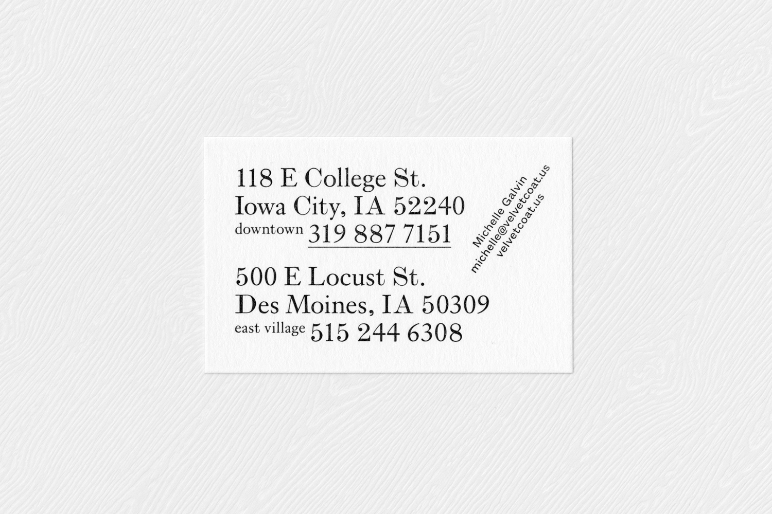 A business card with elegant typography displaying addresses and contact details resting on textured white paper.