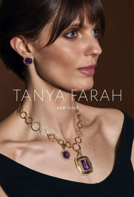 Close-up of an adult woman wearing elegant jewelry, including a gold necklace and amethyst earrings, against a dark neutral background.