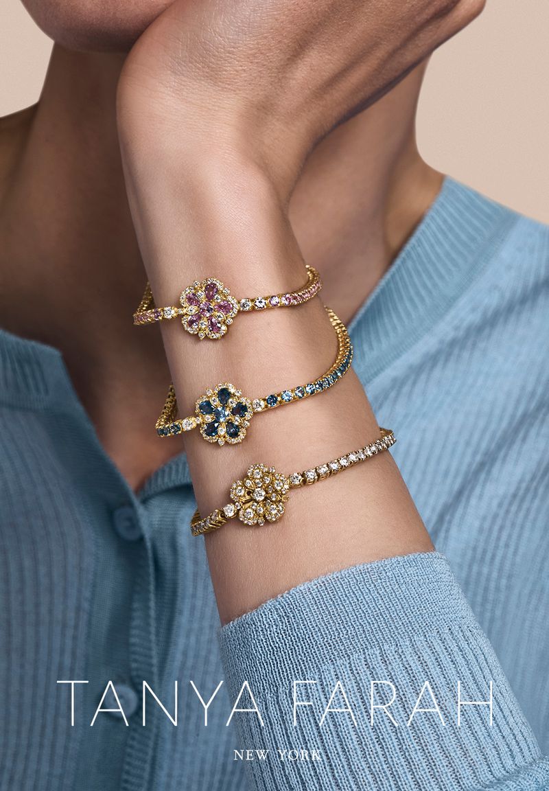 Wrist adorned with three gold flower bracelets featuring colorful gemstone petals, set against a soft blue long-sleeved sweater backdrop.