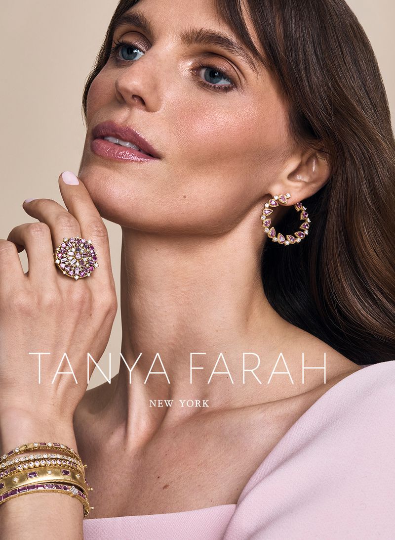 Close-up of a woman modeling fine jewelry including hoop earrings, a cocktail ring, and bangles, against a neutral backdrop.