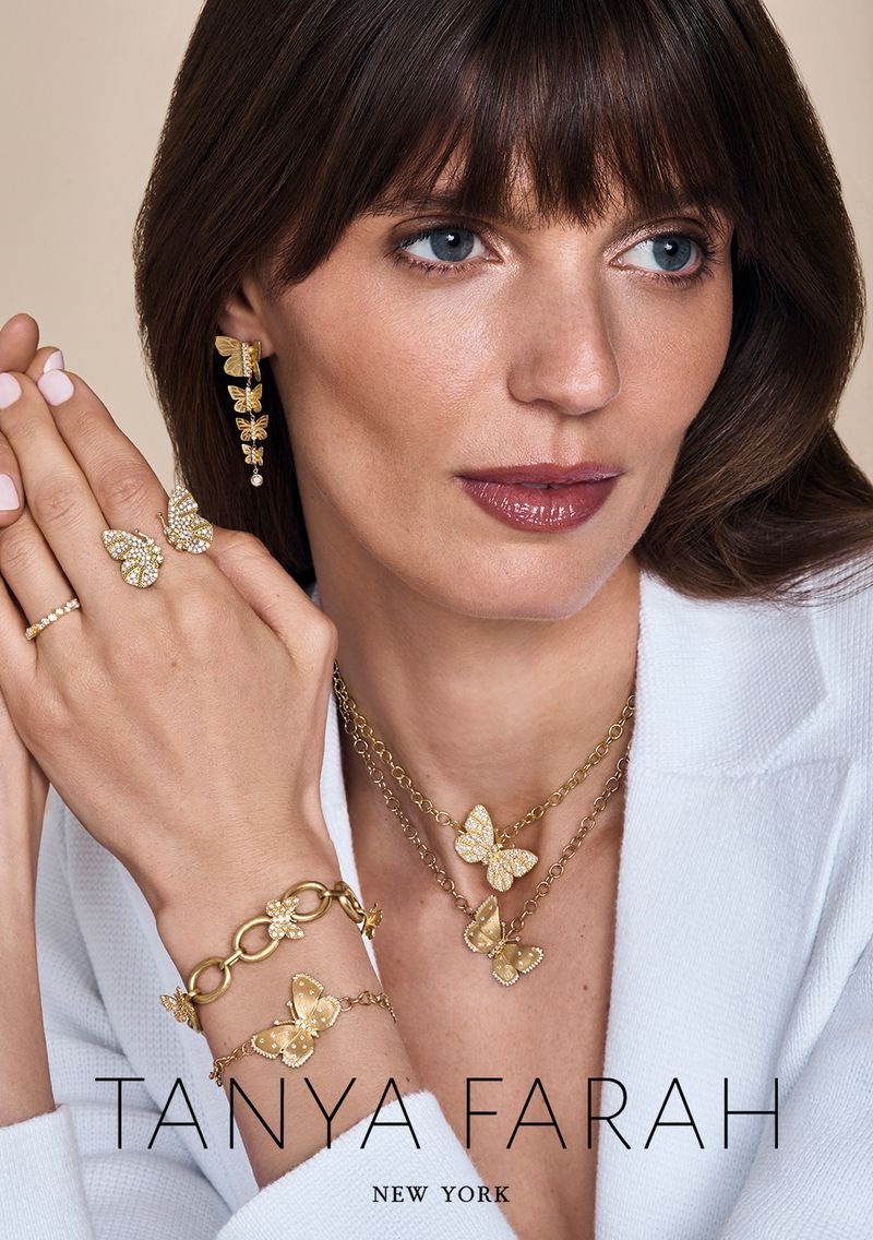 Close-up of an adult woman wearing gold butterfly jewelry, including earrings, necklaces, a ring, and a bracelet, against a neutral backdrop.