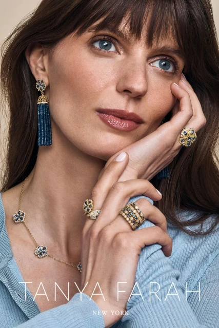 Close-up of a brunette woman with blue eyes wearing a light blue sweater and an assortment of gold and blue jewelry.