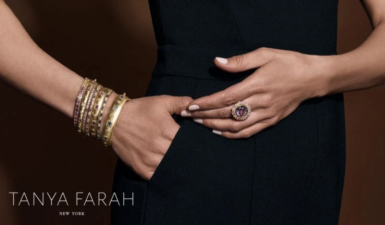 Close-up of a woman wearing gold bracelets and a ring, showcasing fashion jewelry against a dark brown background. Jewelry advertisement.