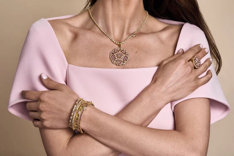 Woman showcases a luxurious jewelry set with a necklace, ring, and bracelets against a neutral backdrop, highlighted by pink dress.
