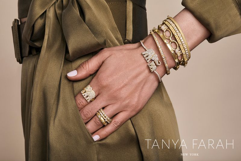 Close-up of a hand adorned with gold and diamond jewelry, including bracelets and rings. An olive green garment is visible.