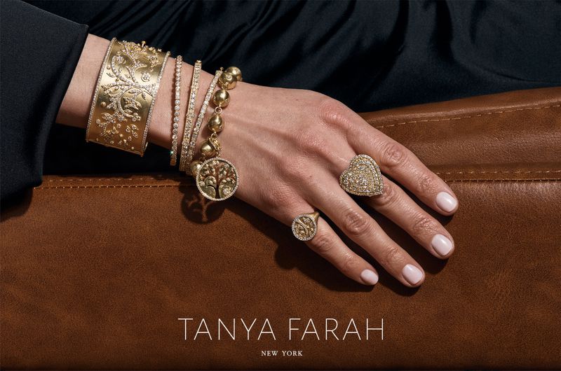 Close-up studio shot showcasing golden bracelets and rings on a woman's hand against a brown textured surface and black fabric backdrop.