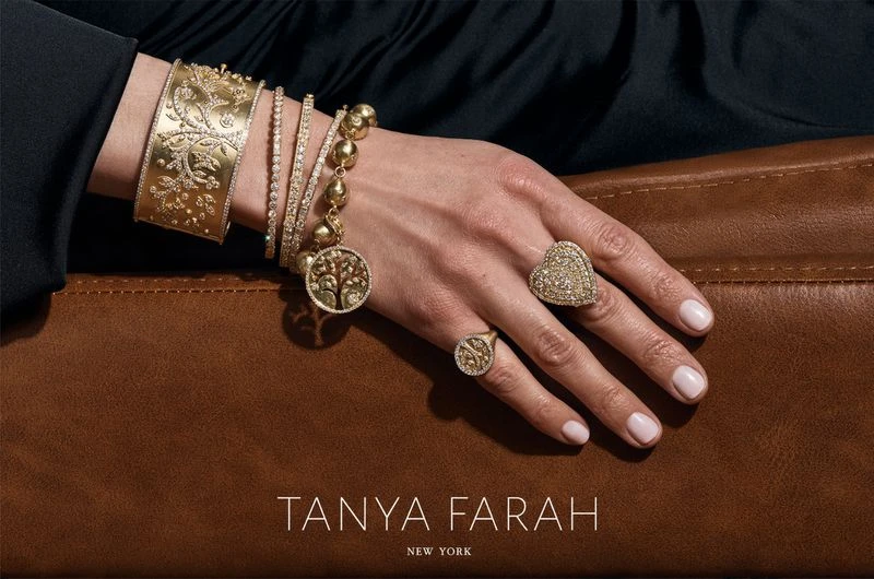 Close-up studio shot showcasing golden bracelets and rings on a woman's hand against a brown textured surface and black fabric backdrop.