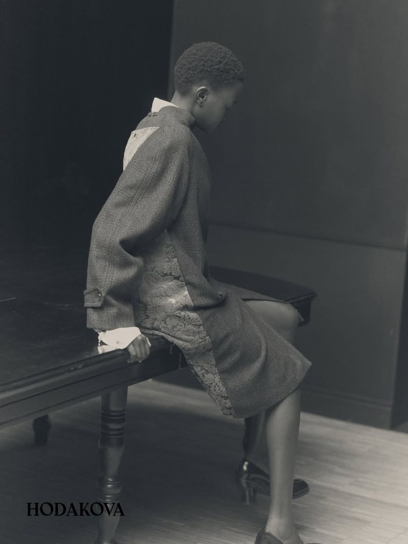 Model wearing a grey textured coat seated on a dark table in a sparse room