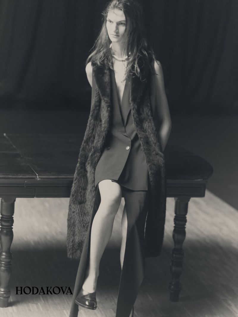 Model seated on a dark wooden table wearing a sleeveless dress and long fur vest for Hodakova