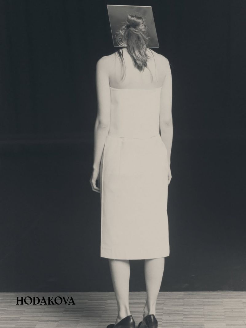 Model from behind wearing a white dress and black shoes with a square prop behind head against dark background.