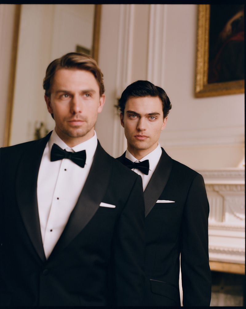 Two men in classic black tuxedos and bowties posing indoors in front of a fireplace mantel and framed painting.