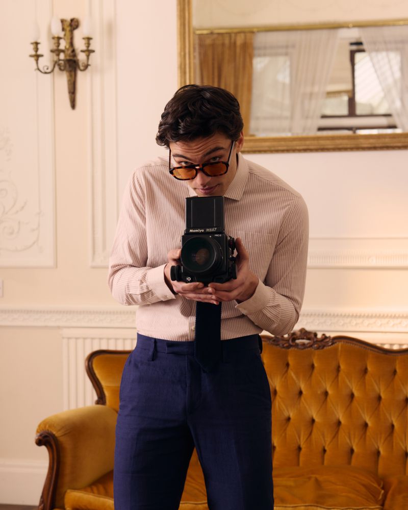 Man wearing a striped shirt and blue trousers holding a vintage medium format camera in an interior room