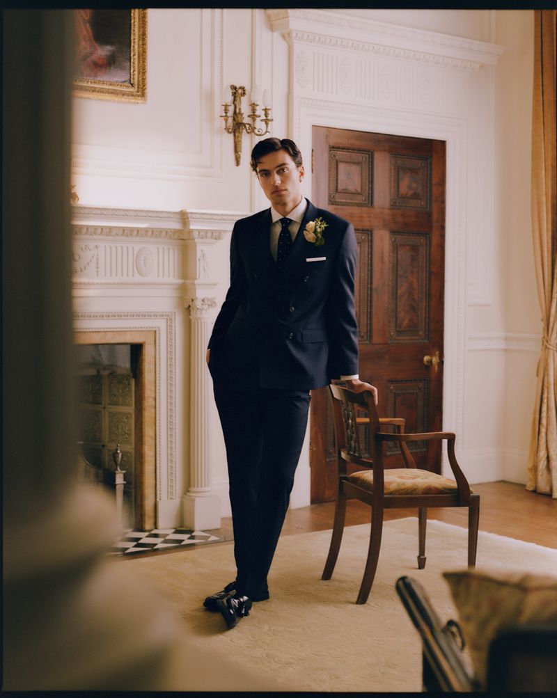 Man in navy blue suit resting hand on wooden chair in an ornate room with a fireplace and wood door
