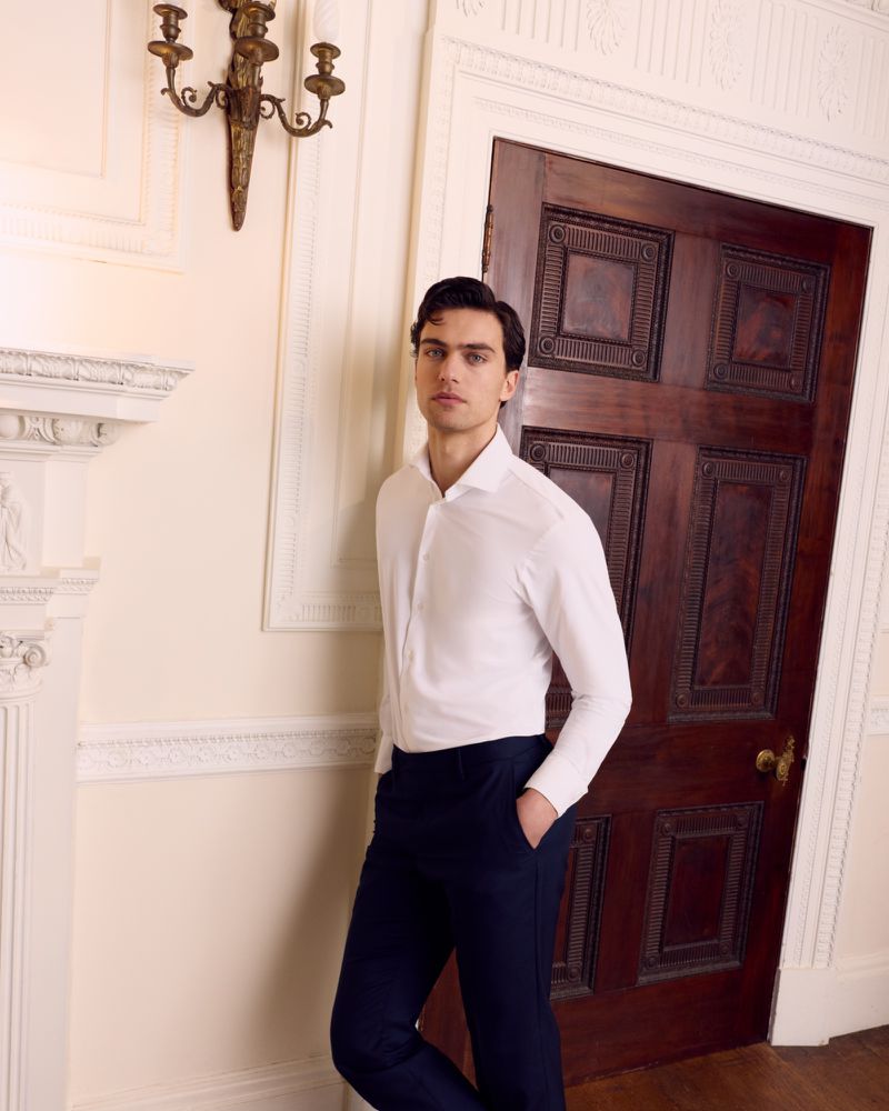 A man wearing a white dress shirt and dark trousers standing indoors by a dark wooden door and ornate interior molding.