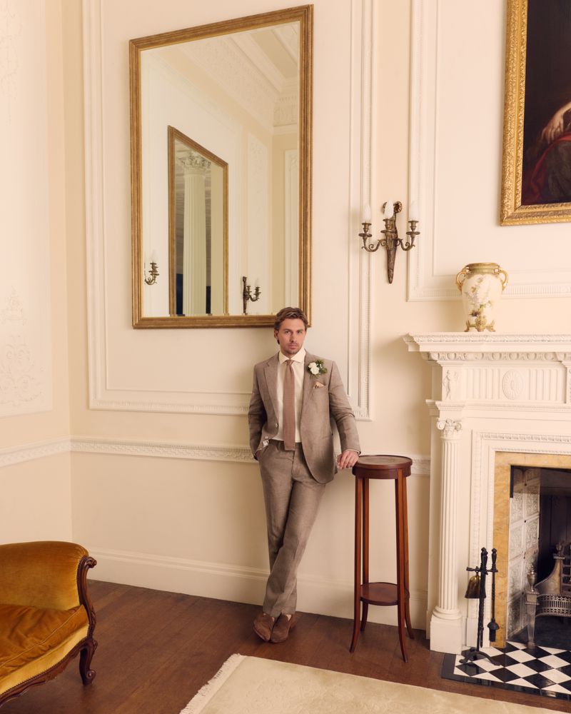 Man in a tan suit leaning against a mantelpiece in an elegant room with a large mirror by Stewart Bryden