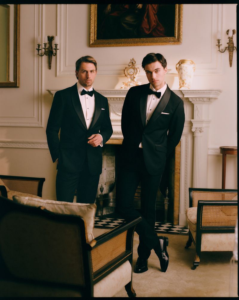 Two men in classic black tuxedos with bow ties standing inside an ornate living room with a fireplace and armchairs.