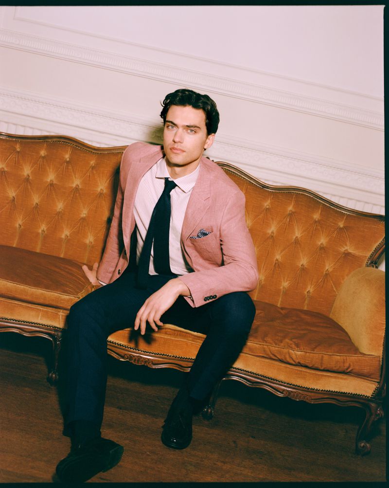A man in a pink blazer and dark trousers seated on a vintage tufted sofa, photographed by Stewart Bryden.
