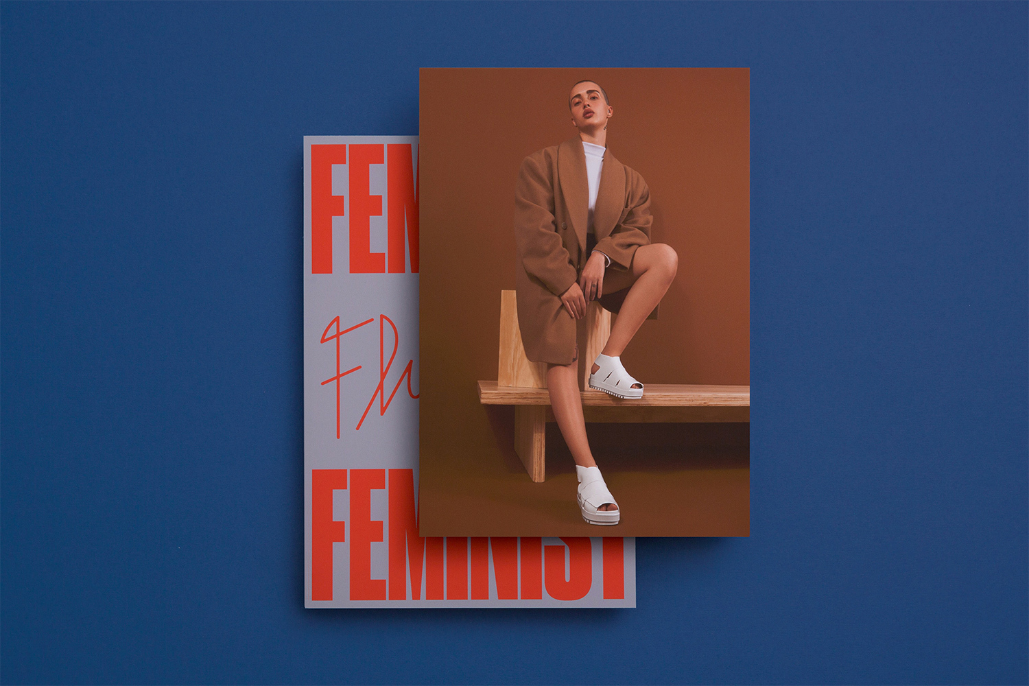 Model wearing a brown coat and white platform shoes seated on a wooden bench with printed promotional materials against a solid blue wall.