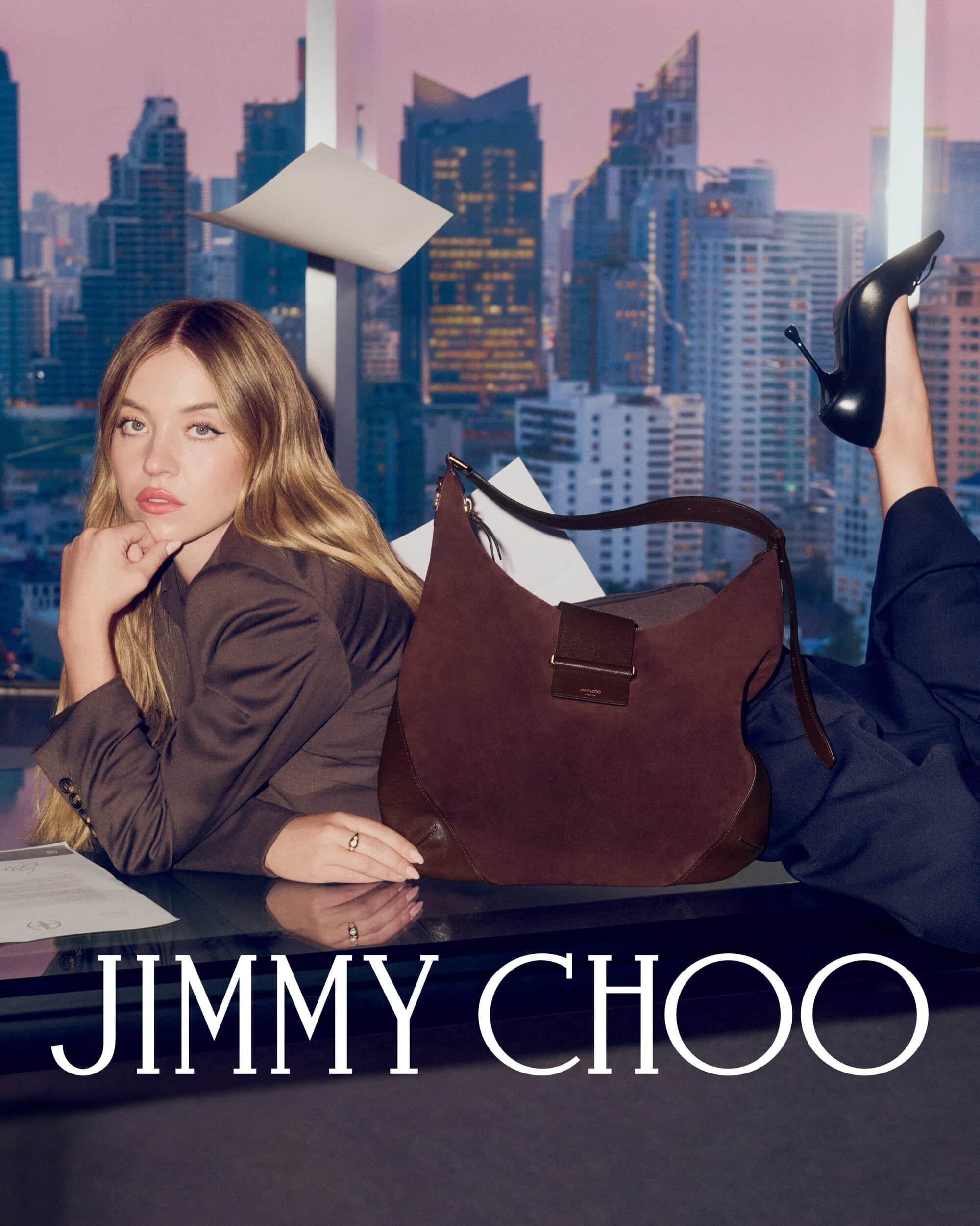 Sydney Sweeney reclining with a brown suede shoulder bag and black stiletto heel against a city skyline background.