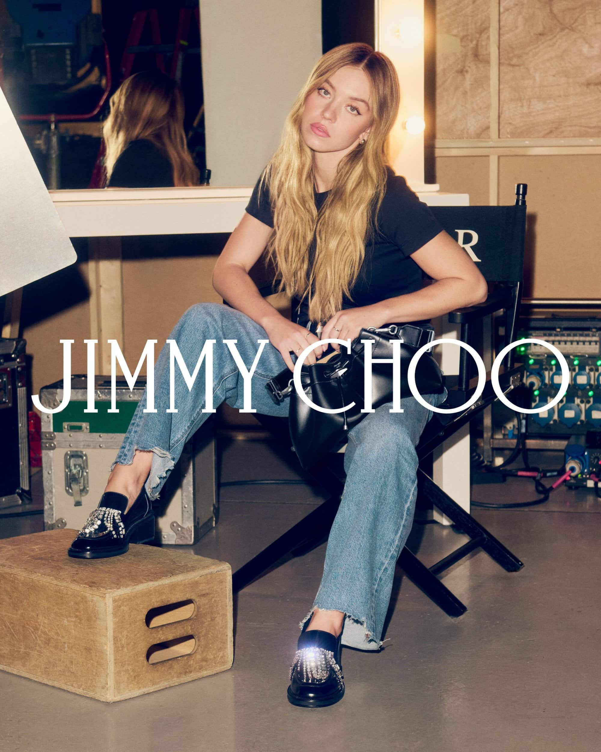 Sydney Sweeney seated in a director's chair wearing a black t-shirt, jeans, and crystal-embellished loafers holding a black handbag.