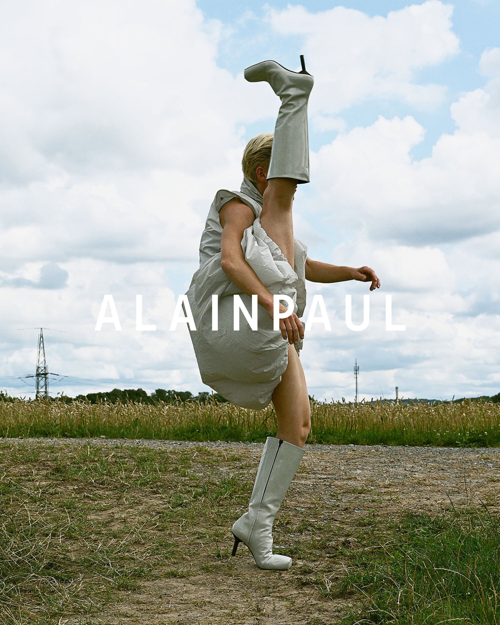 Model with high-extended leg wearing white boots and dress against an open field with cloudy sky.