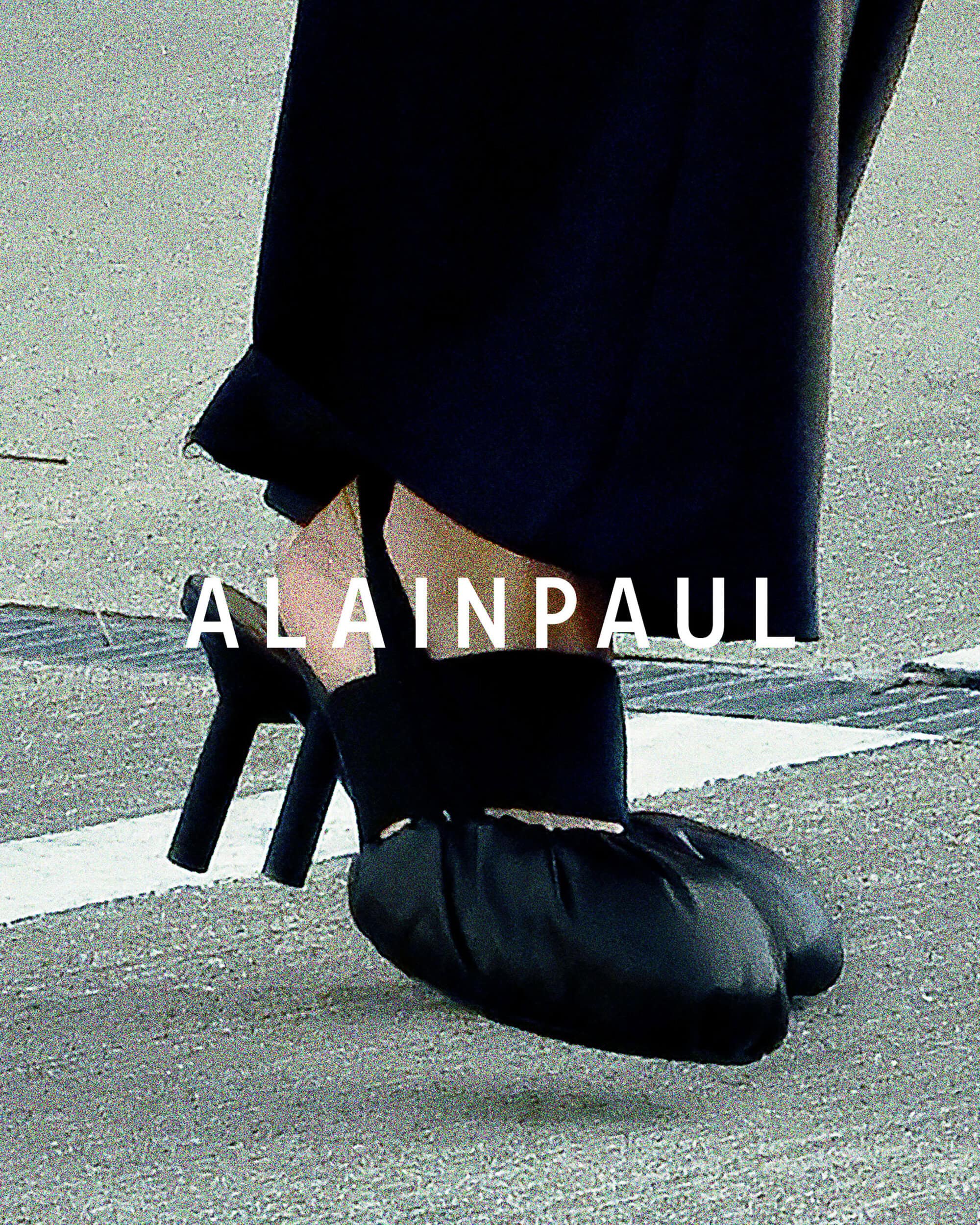 Close-up of a person wearing black heeled sandals with gathered fabric toes and dark trousers over a textured concrete surface.