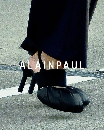 Close-up of a person wearing black heeled sandals with gathered fabric toes and dark trousers over a textured concrete surface.