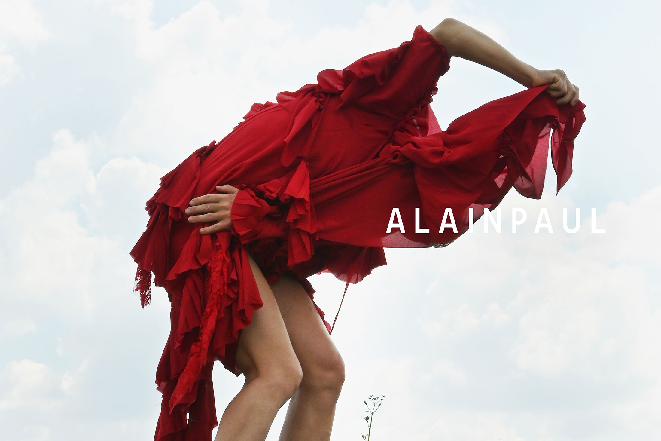 Model in a draped red dress bending forward against an overcast sky for Alainpaul