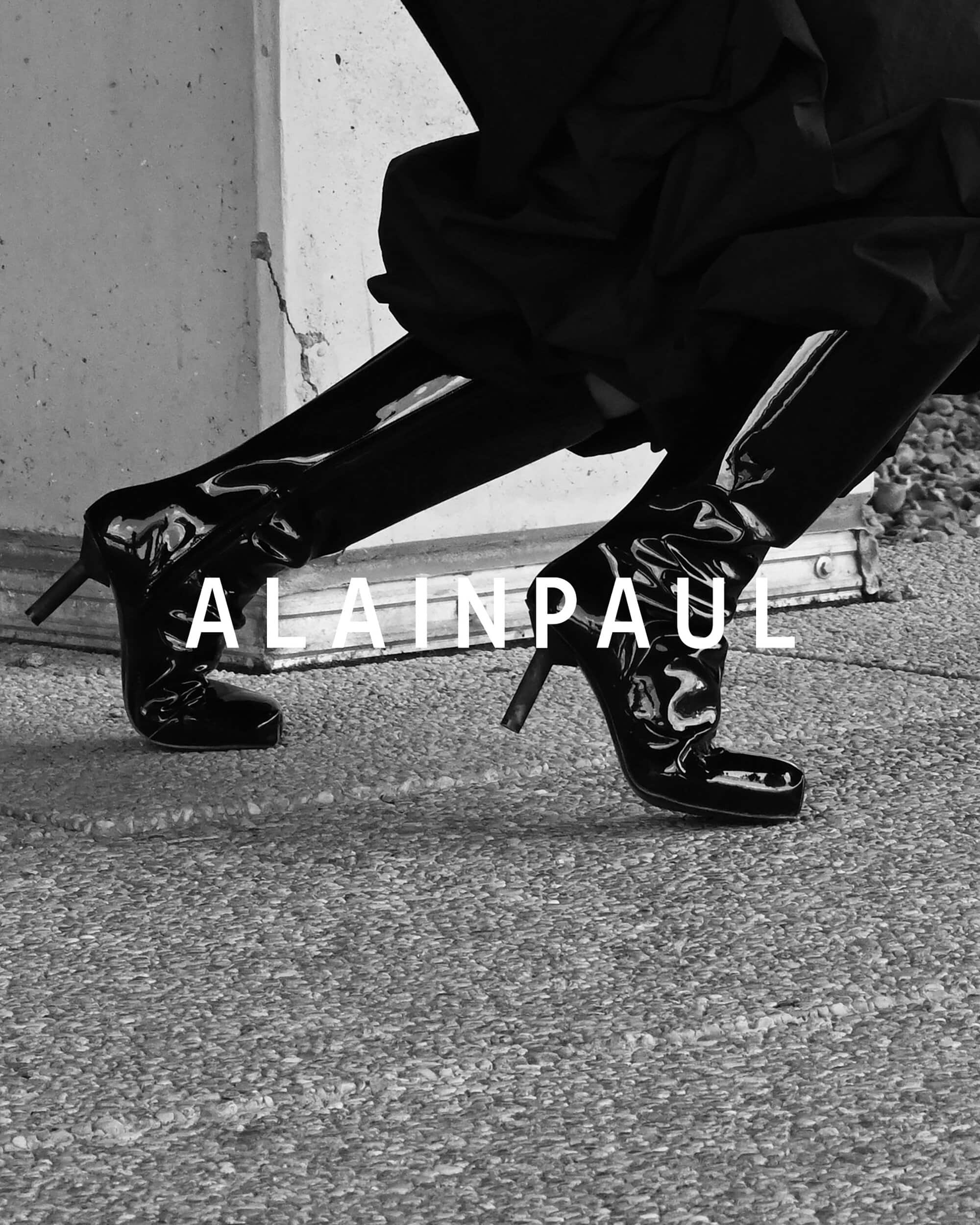 Close-up of a person wearing glossy black patent leather boots and dark apparel outdoors against a textured concrete wall.