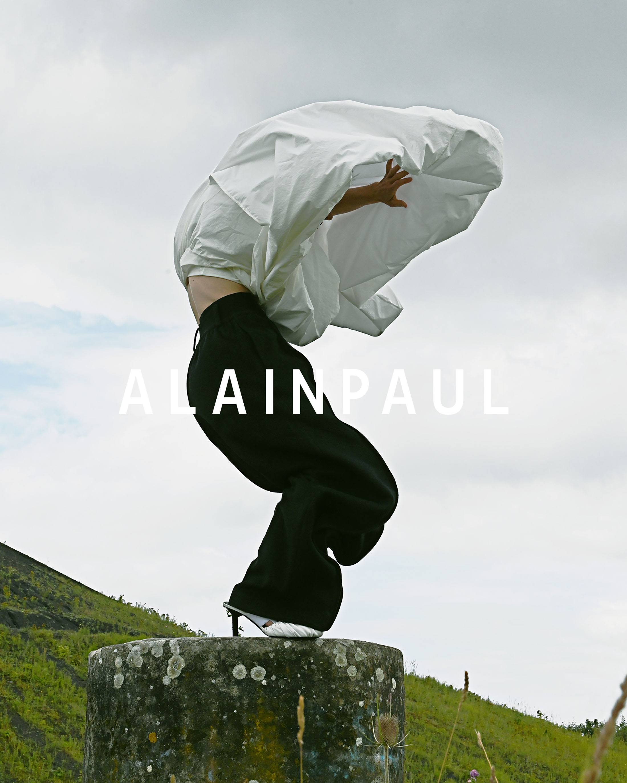 Model balancing acrobatically on a concrete pillar outdoors wearing black trousers and holding white fabric under a cloudy sky