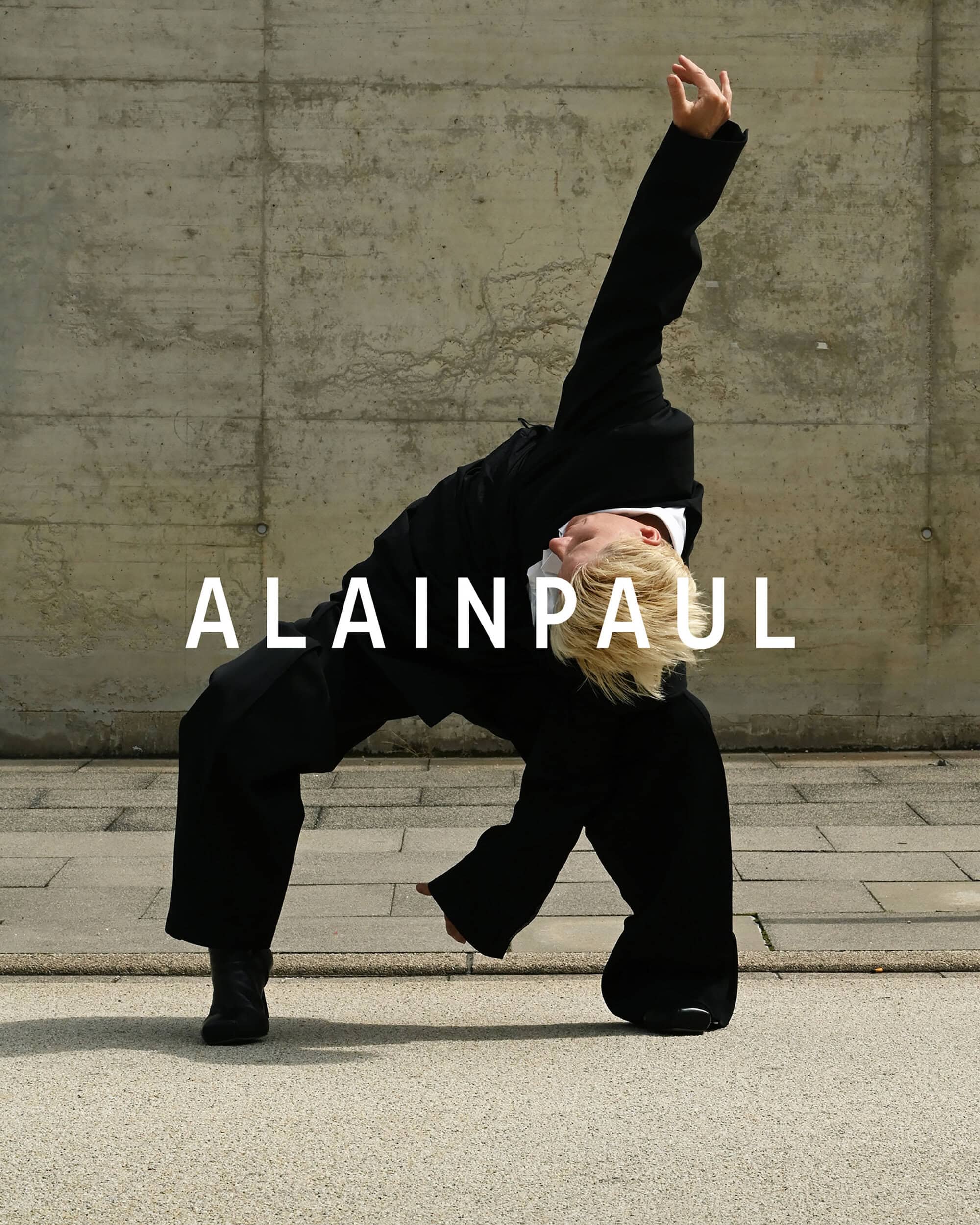 Model in black suit leaning backward against a concrete wall for Alainpaul ad campaign