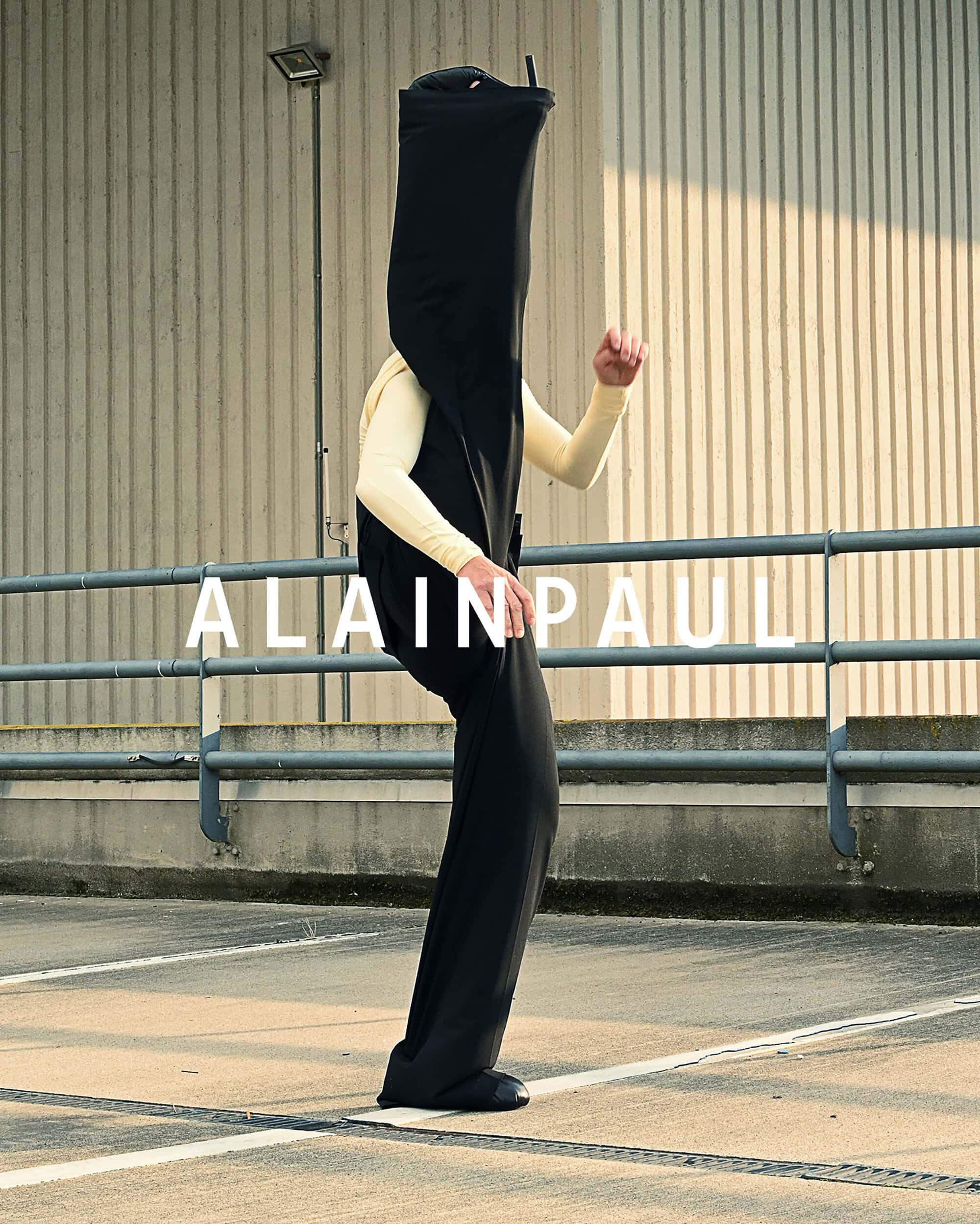 Model wearing a black garment obscuring their head against a corrugated metal wall and concrete ground