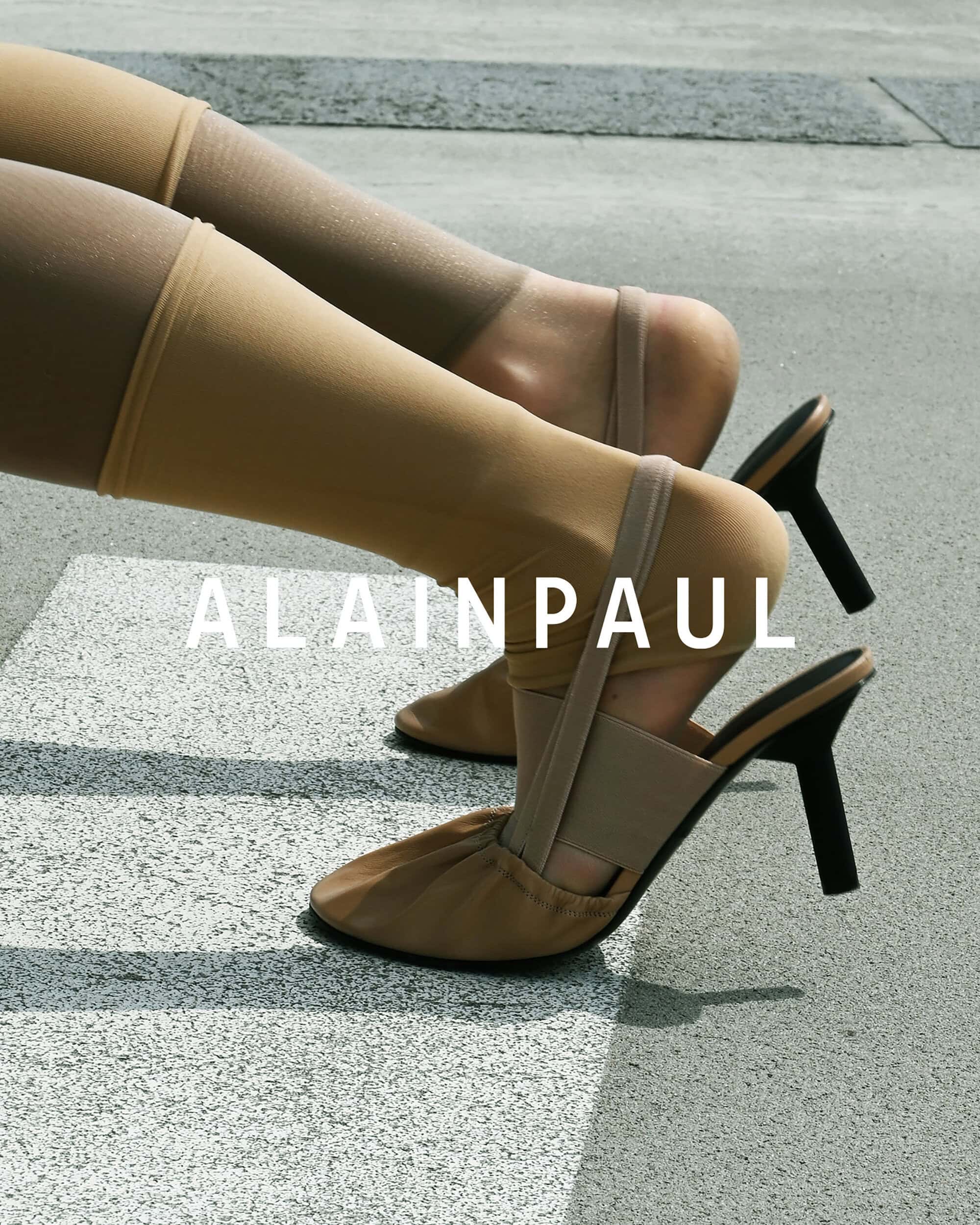 Close-up of legs wearing nude stockings and heeled mules on outdoor pavement with the Alainpaul brand logo overlay