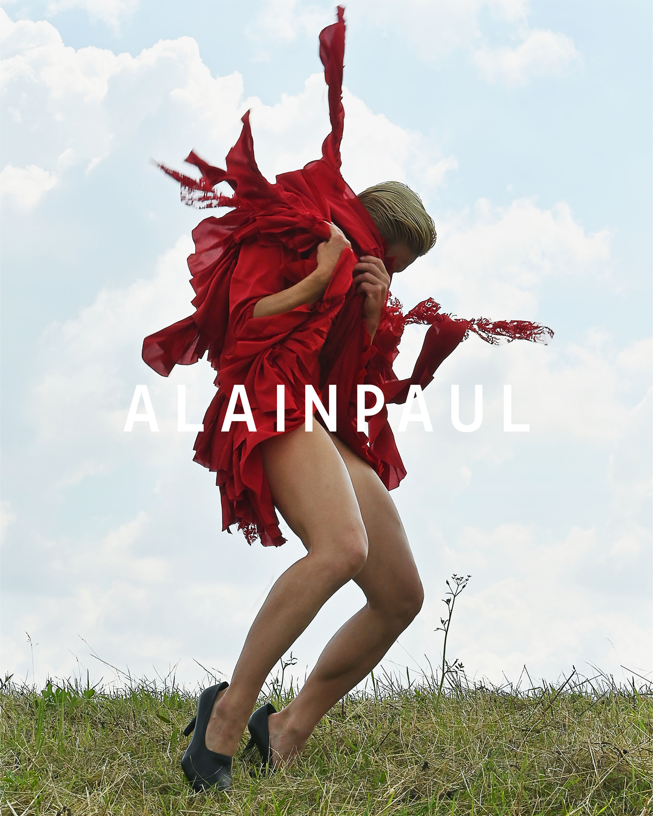 Model wearing a vibrant red dress caught mid-motion outdoors against a cloudy sky with grassy foreground