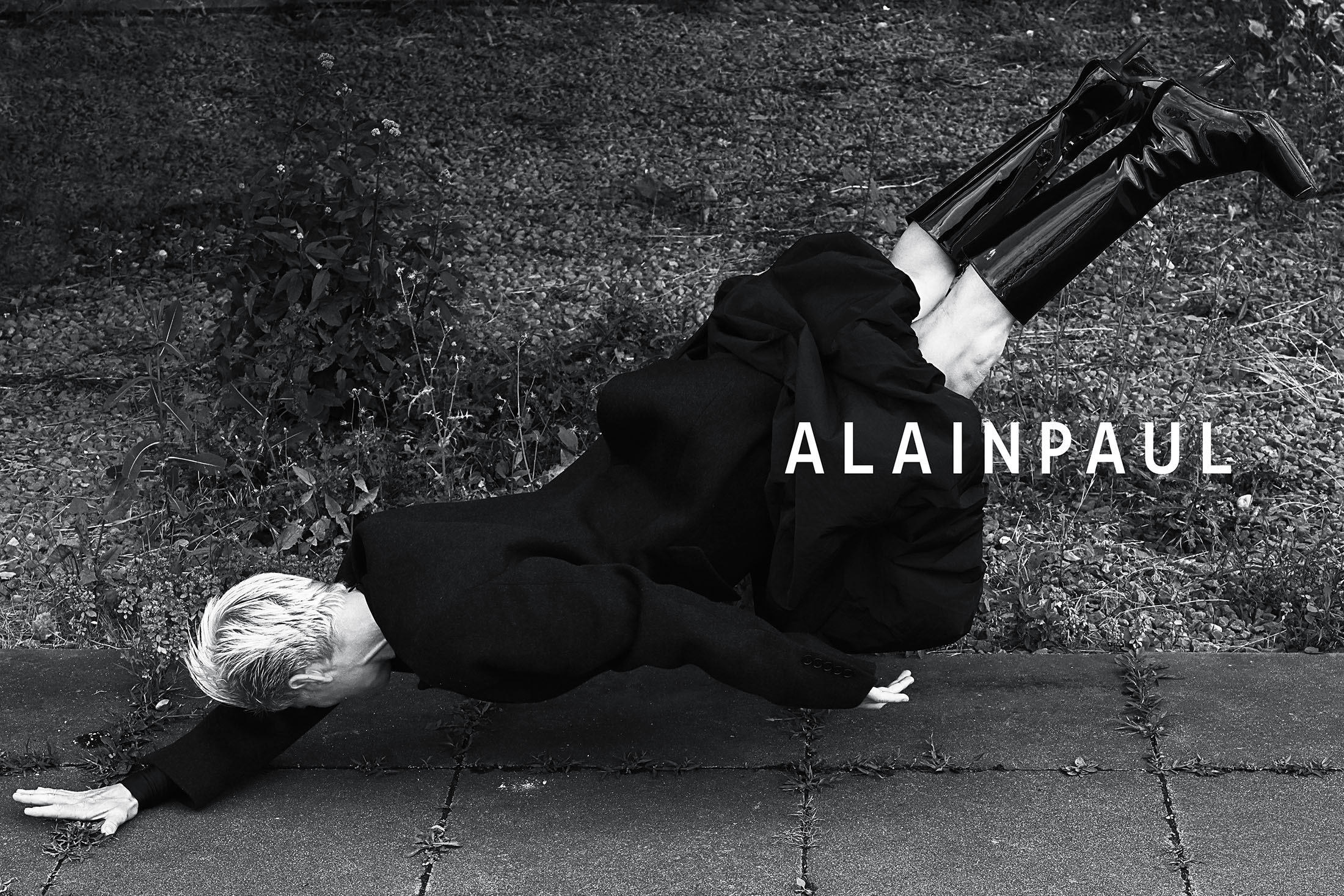 Model wearing a black coat and patent leather boots suspended horizontally over paved ground with vegetation.
