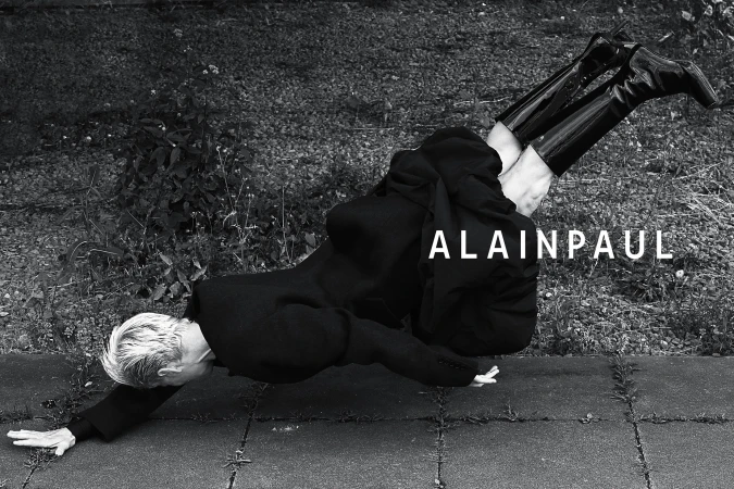 Model wearing a black coat and patent leather boots suspended horizontally over paved ground with vegetation.