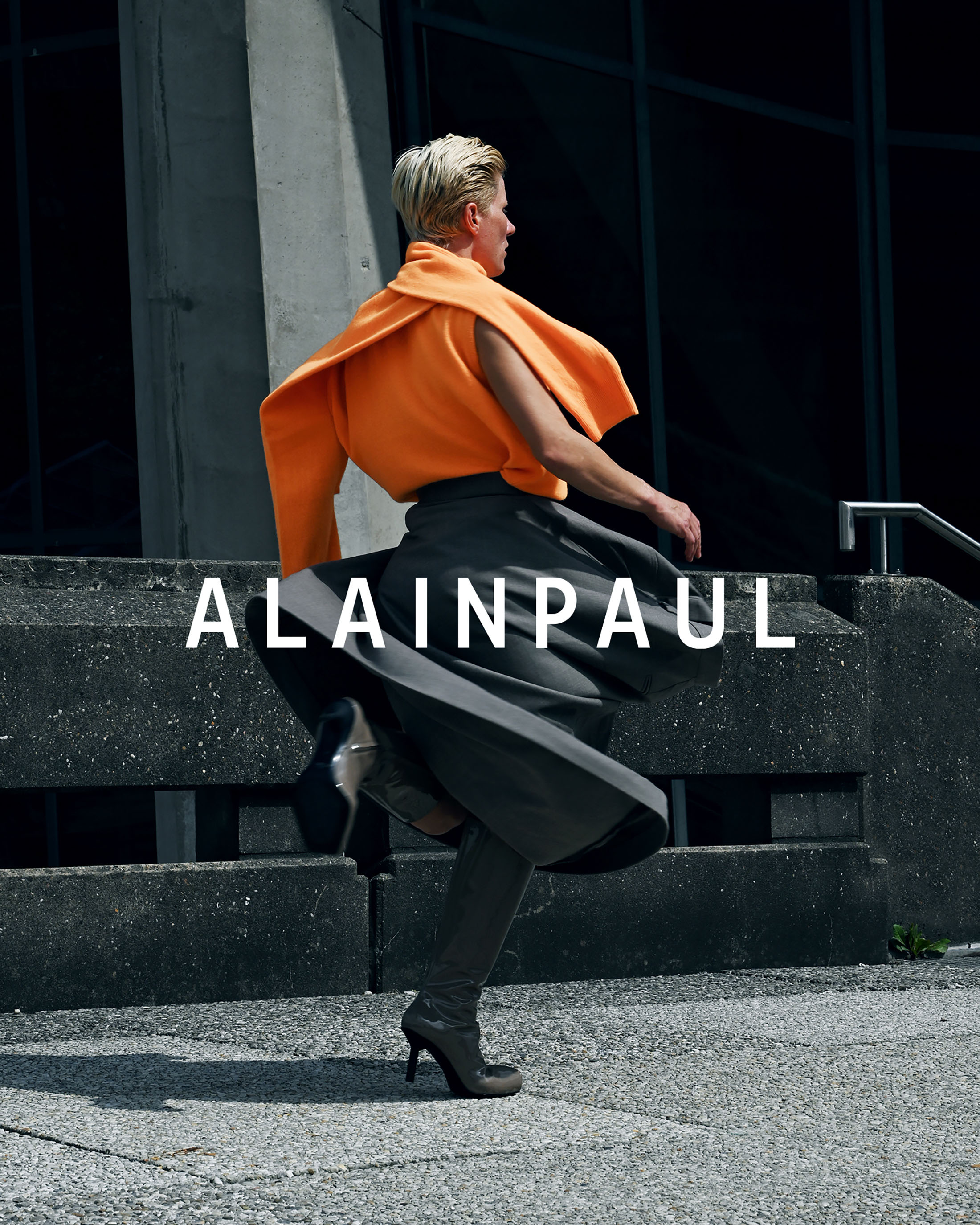 Model with blonde hair wearing an orange top and grey skirt posing dynamically outside concrete architecture