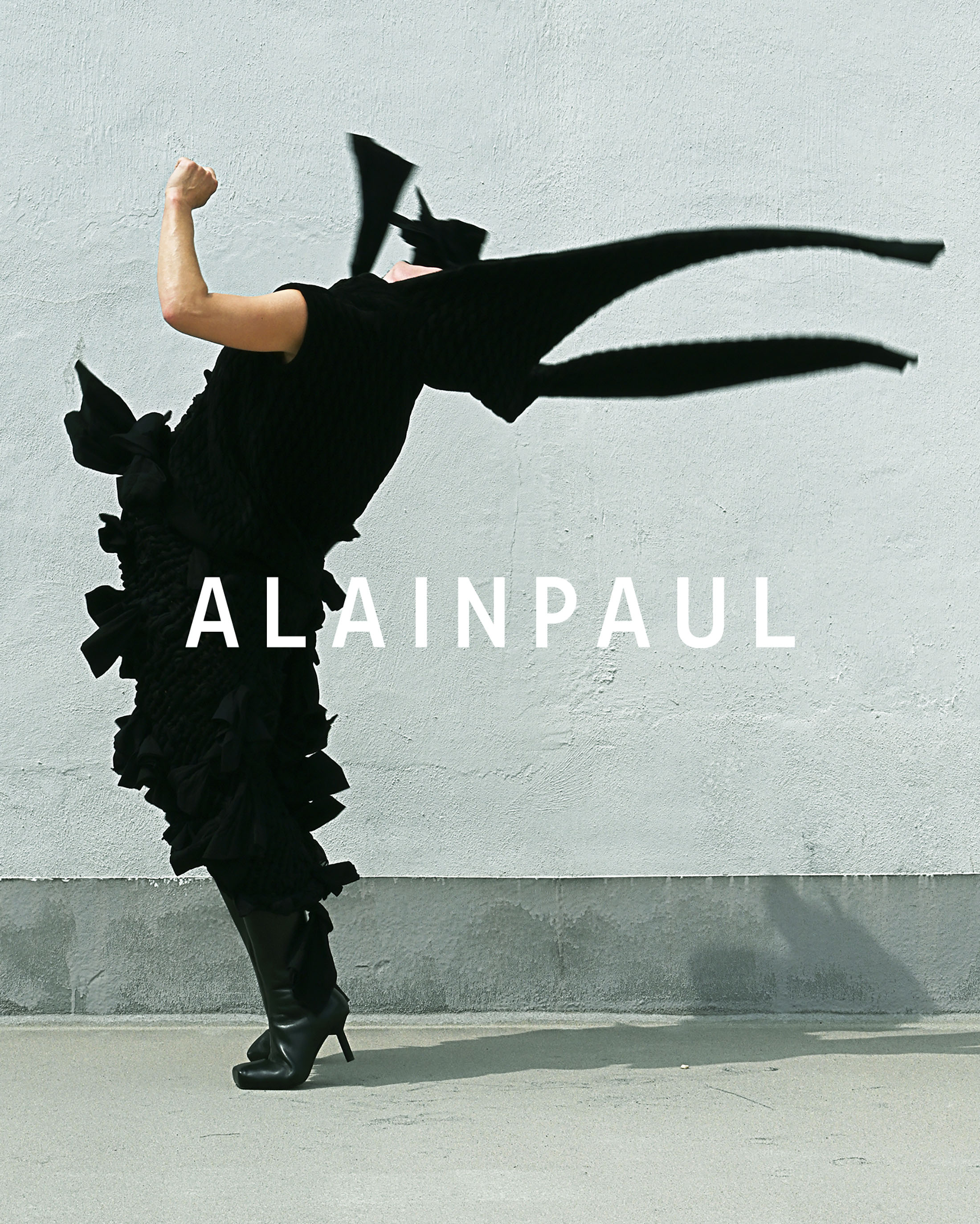 Model in dynamic arched pose wearing black sculptural dress against a textured concrete wall with Alainpaul branding