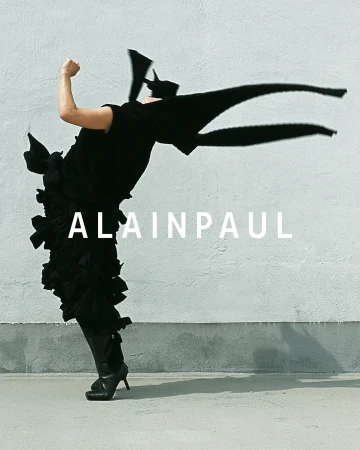 Model in dynamic arched pose wearing black sculptural dress against a textured concrete wall with Alainpaul branding