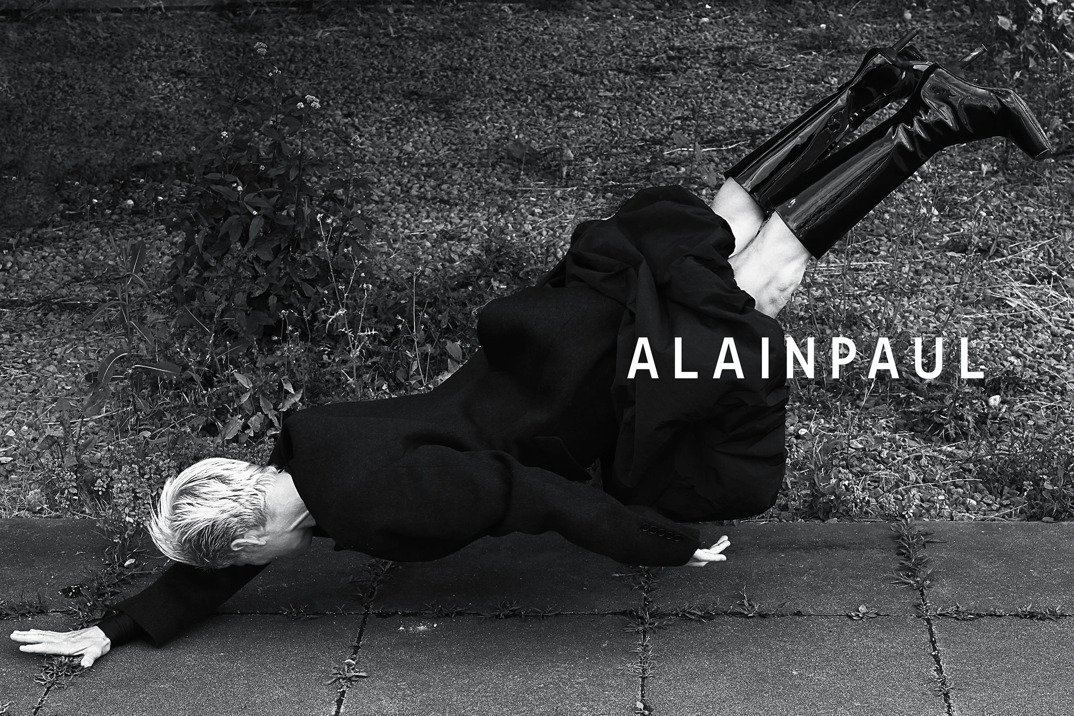 Model wearing a black coat and patent leather boots suspended horizontally over paved ground with vegetation.