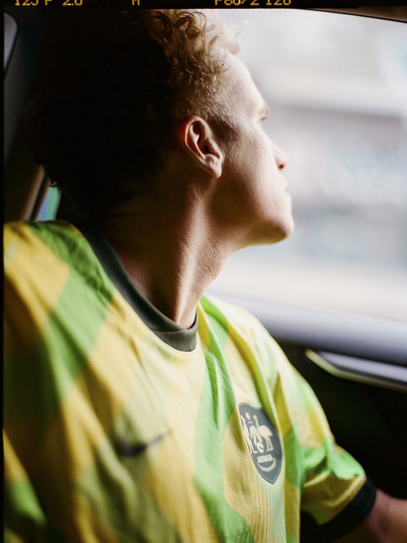 Man in green and yellow soccer jersey looks out car window with sunlit hair, capturing a candid portrait.