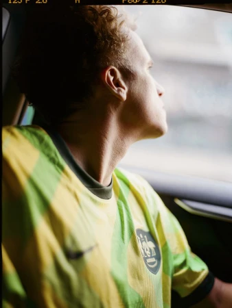 Man in green and yellow soccer jersey looks out car window with sunlit hair, capturing a candid portrait.