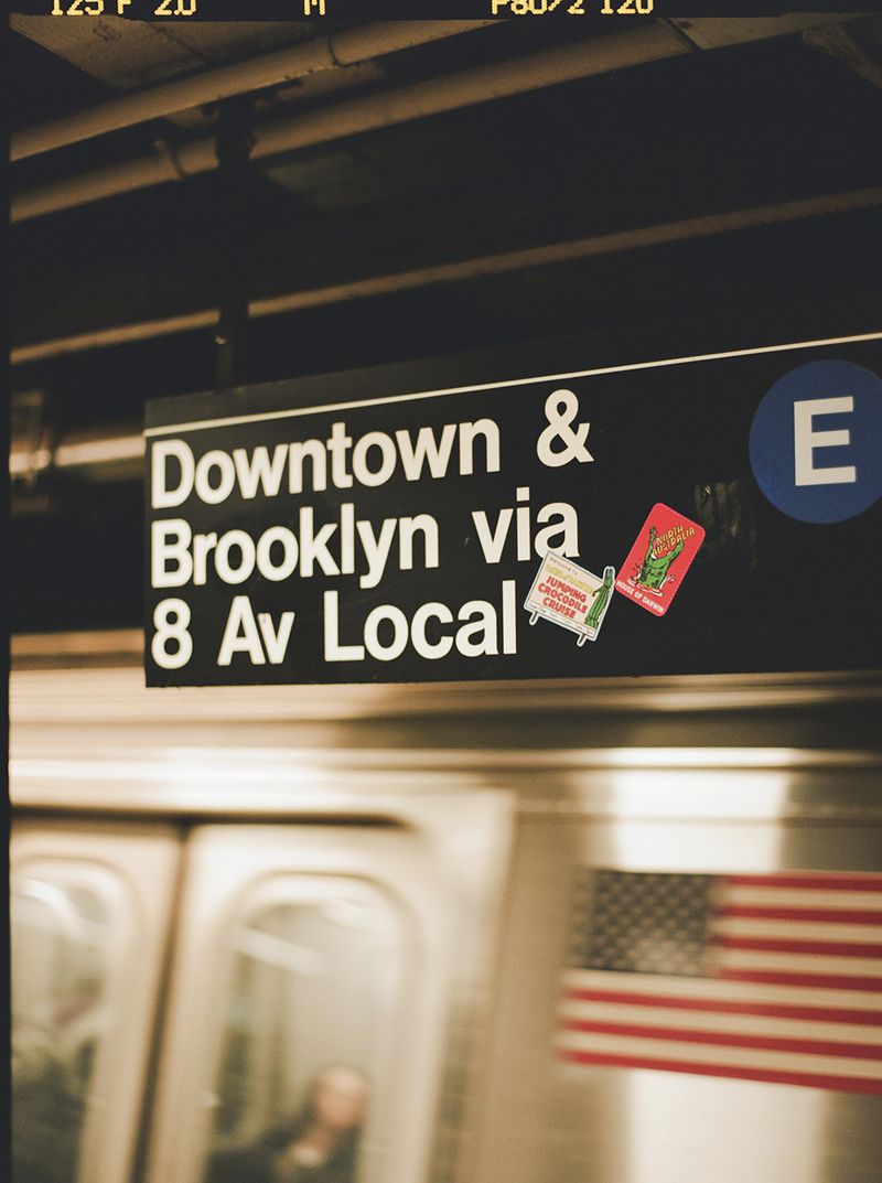Subway platform sign for Downtown & Brooklyn via 8 Av Local with stickers, train, and blurred figure, creating an urban scene.