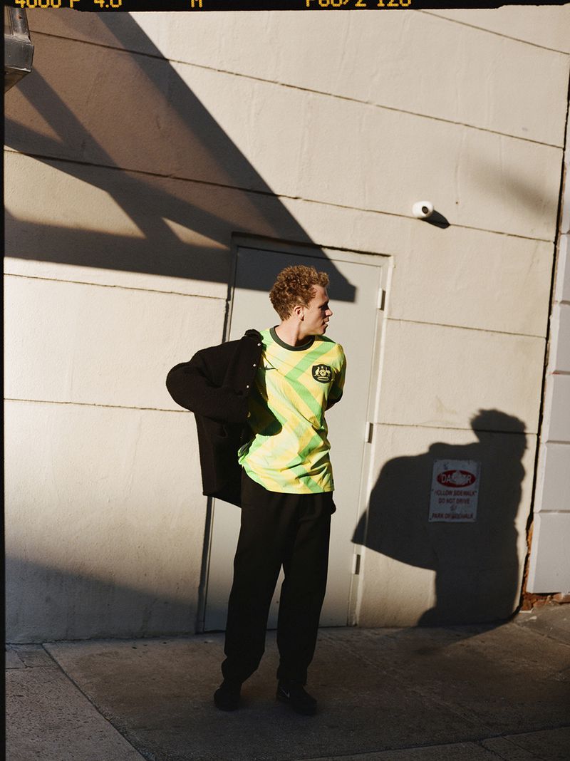 Man wearing a yellow and green striped shirt and black pants poses in front of a white concrete building on a sunny day.