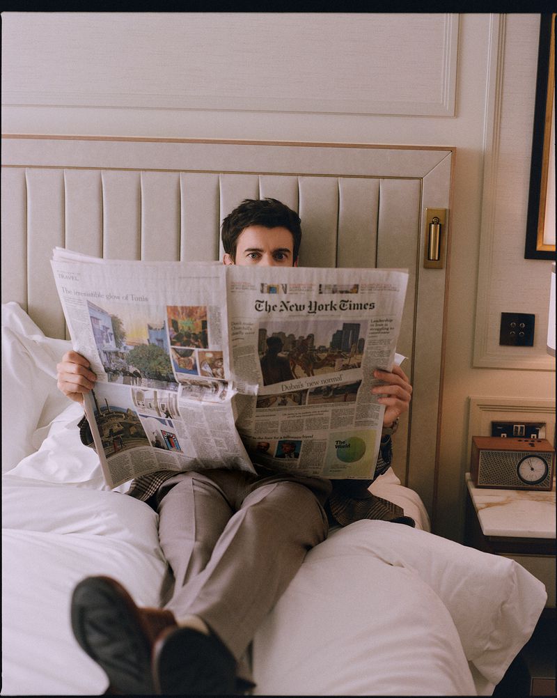 Man sitting on a hotel bed holding a large newspaper that hides most of his face, photographed by Stewart Bryden.