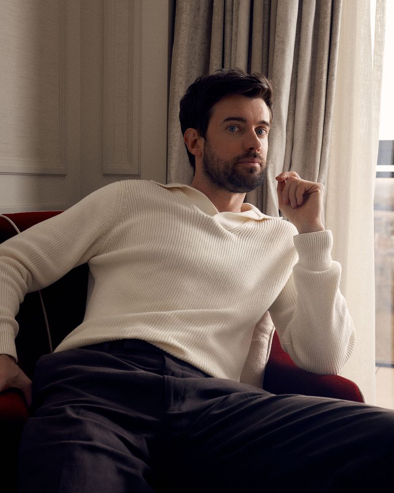 Jack Whitehall seated in a red armchair wearing a cream polo sweater and dark trousers near a draped window.