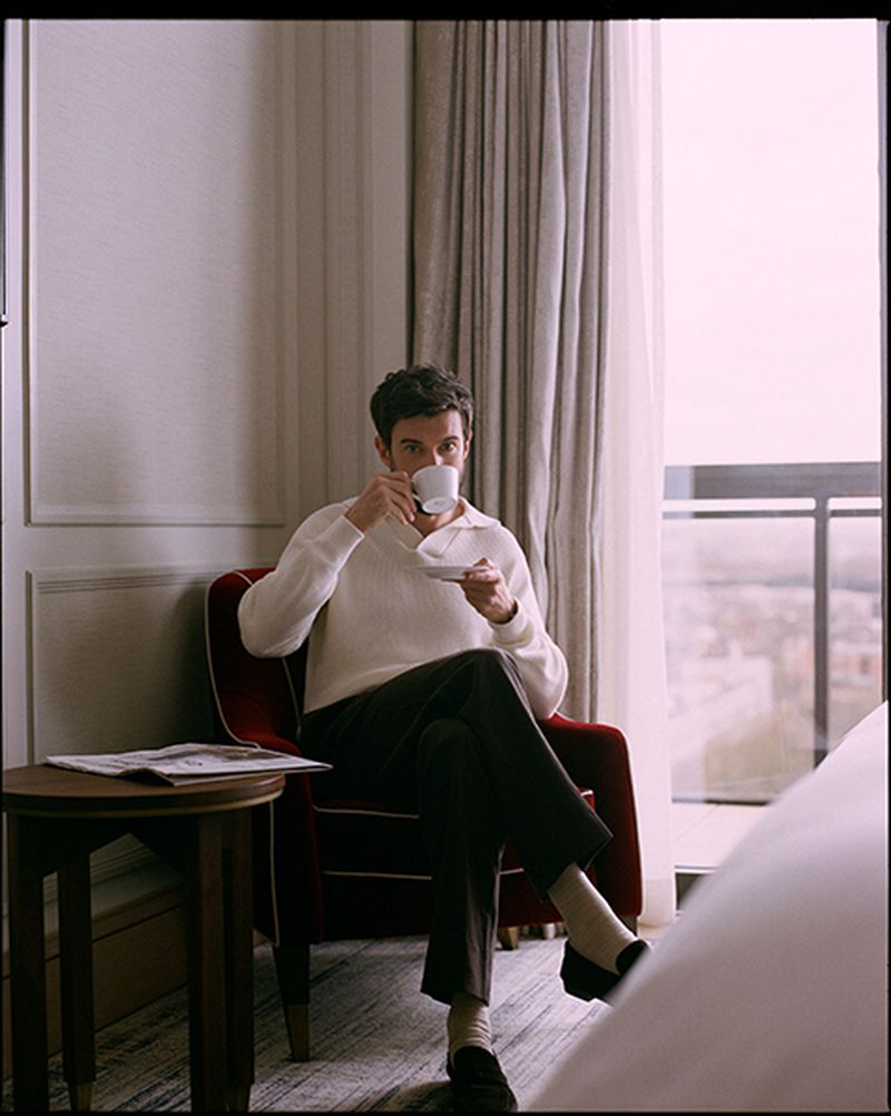 A man seated in a red armchair in a hotel room, drinking from a white teacup while holding a saucer.